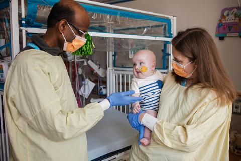 A masked doctor gestures to a baby who is being held by another masked doctor.
