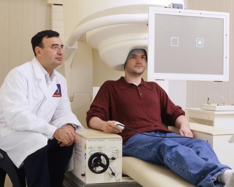 Zarate in white lab coat seated next to man sitting in a MEG scanner