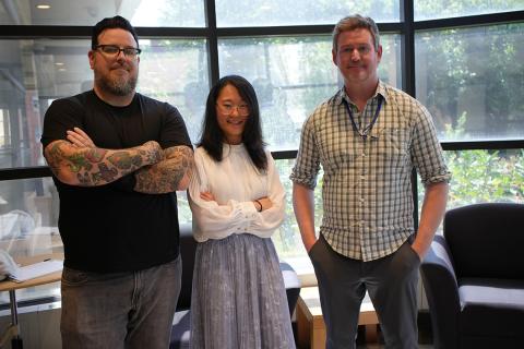 The trio of presenters pose together against a wall of windows.