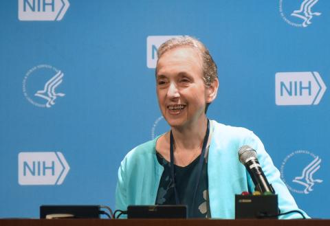 Schor speaks into a microphone. She is wearing a teal cardigan and standing against a light blue background patterned with the NIH and HHS logos.