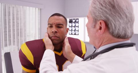 A man in a football jersey seated in a brightly lit room. A doctor with his back to the viewer examines the patient's head. 