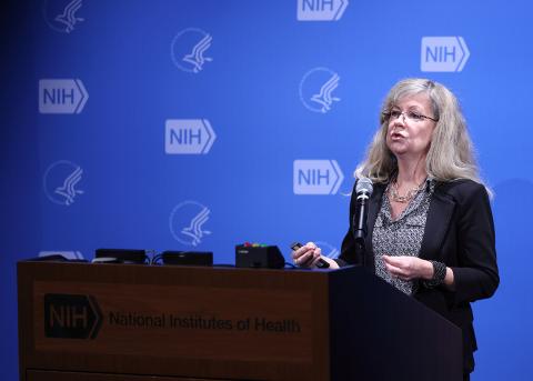 Ten Hagen speaks into a microphone. Behind her is a light blue backdrop featuring NIH and HHS logos.