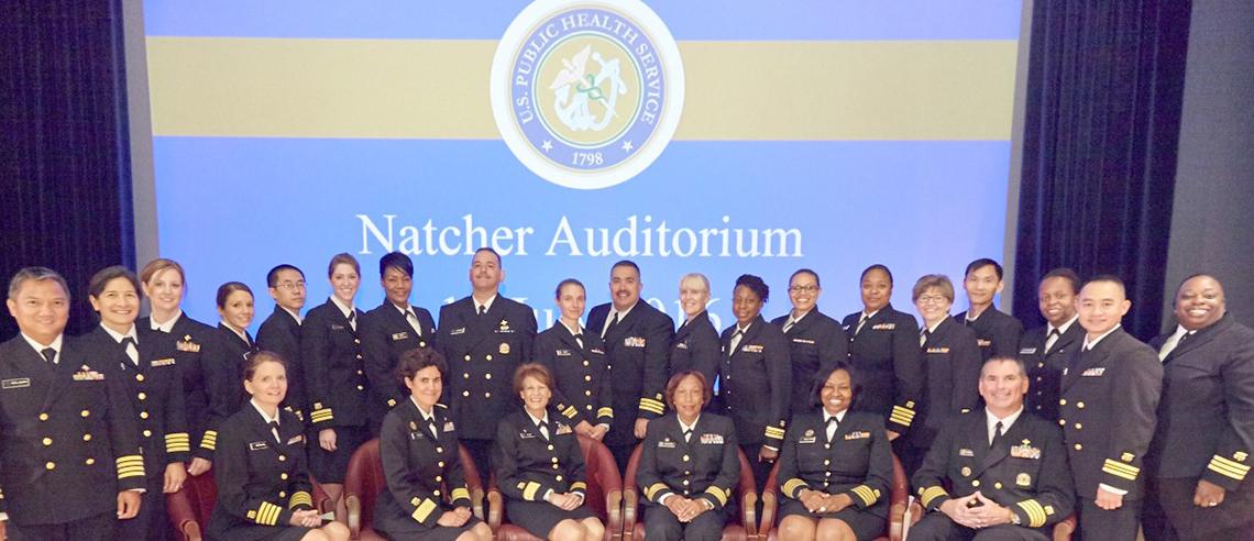 Large group of Commissioned Corps members in front of slide announcing promotion ceremony
