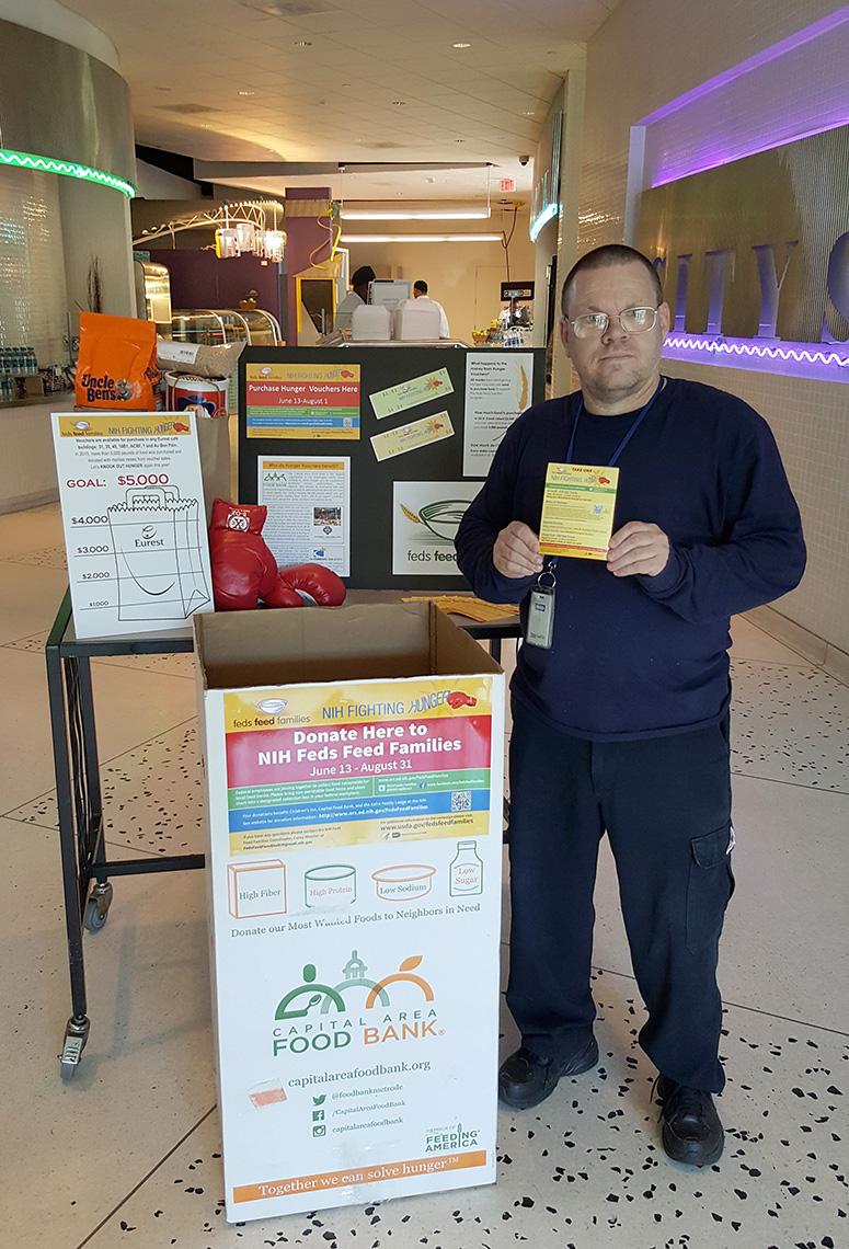 Harrison stands in cafeteria holding hunger-fighting campaign information.