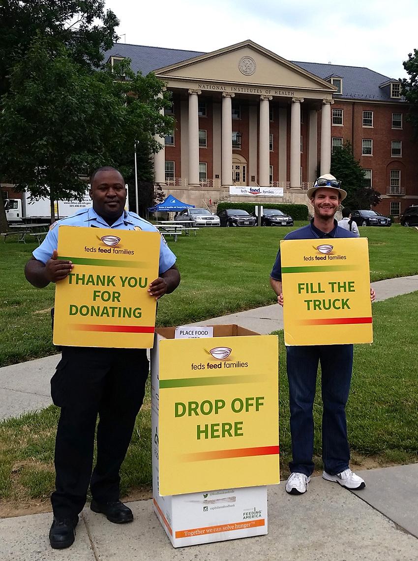 NIH employees hold yellow signs that read: fill the truck, thank you for donating, drop off here in front of Bldg. 1.