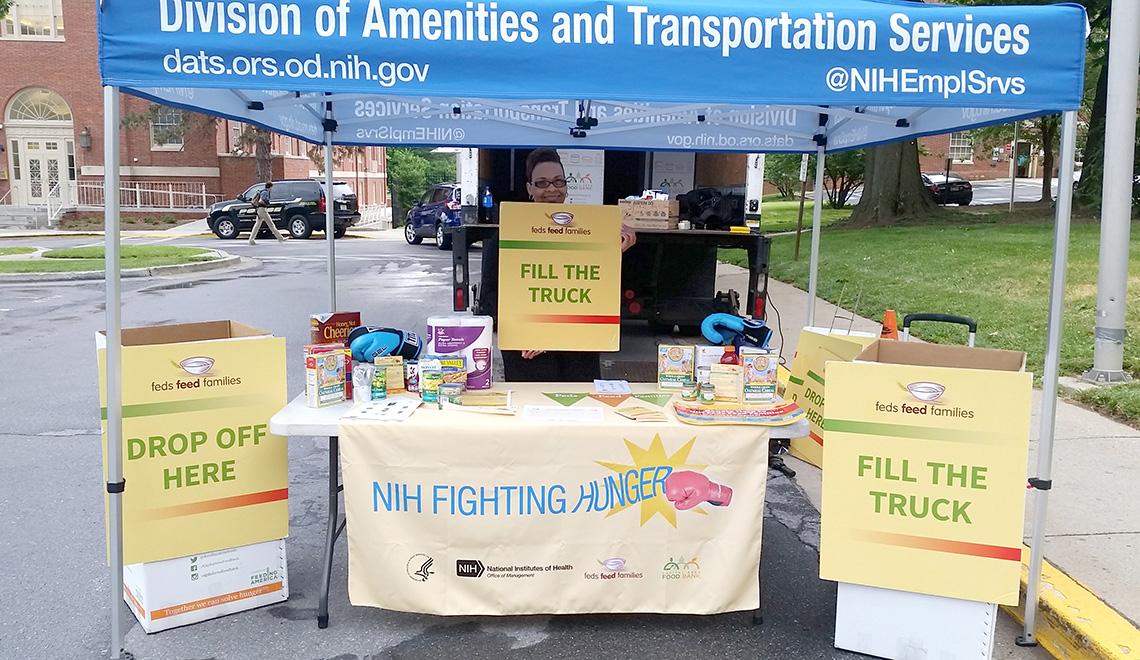 NIH'er stands by drop-off bins under blue DATS tent holding a Fill the Truck sign 