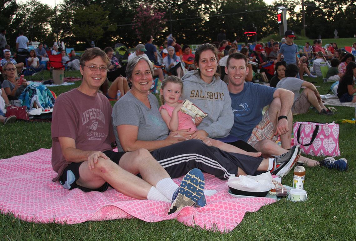 Family sits on pink blanket on the lawn, ready for the movie.