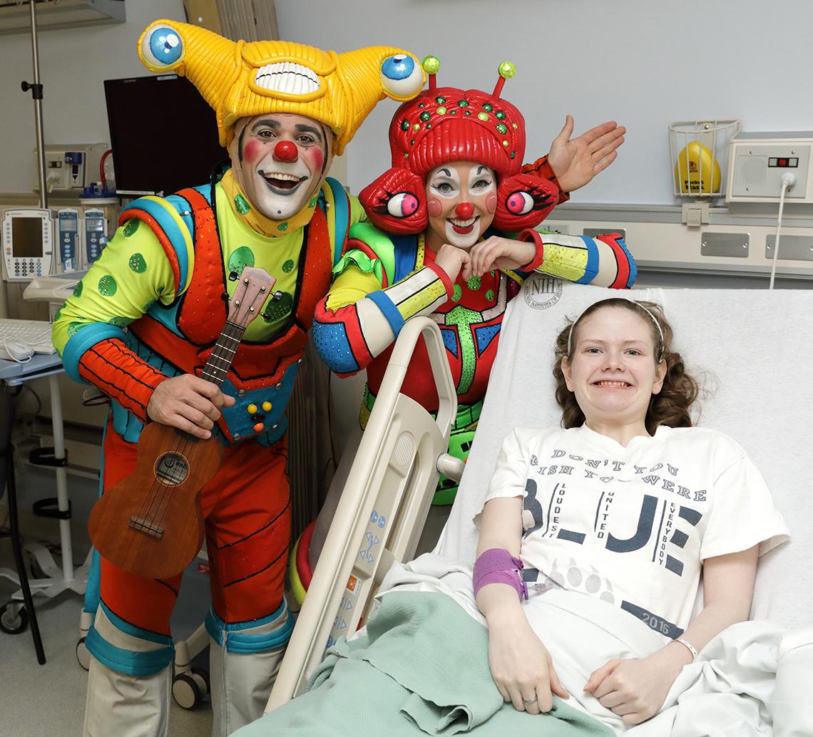 Young girl in hospital bed smiles as clowns pose beside her.