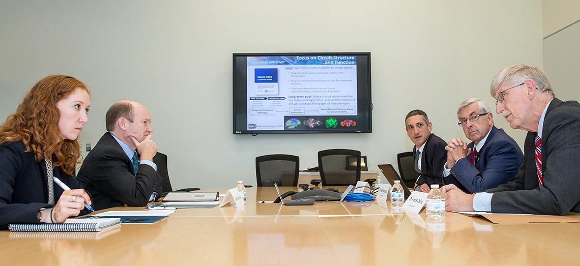 Coons sits across a large table from Reich, Koroshetz and Collins