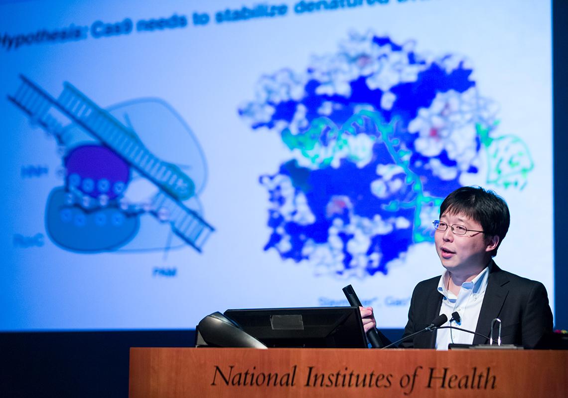Zhang presents at microphone with slide projected behind him.