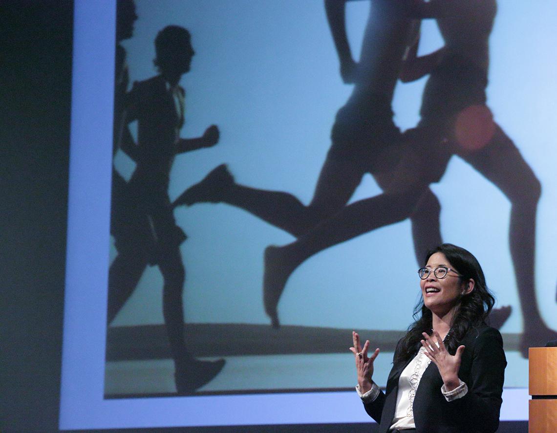 Suzuki holds up hand while speaking, standing in front of slide of joggers