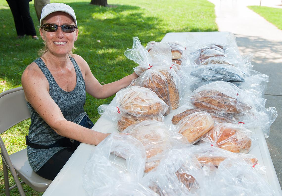 The smiling bread lady sits at table with loaves for sale at outdoor community market.