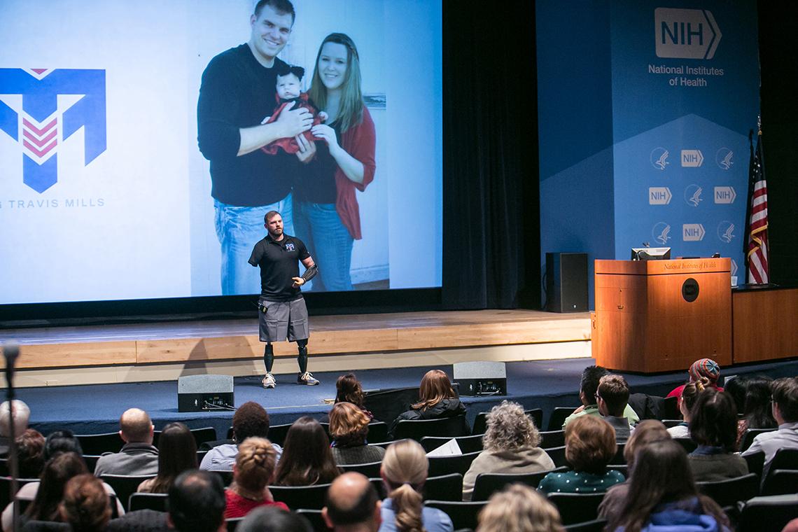 Mills standing on prosthetic legs in front of slide showing his family before his accident