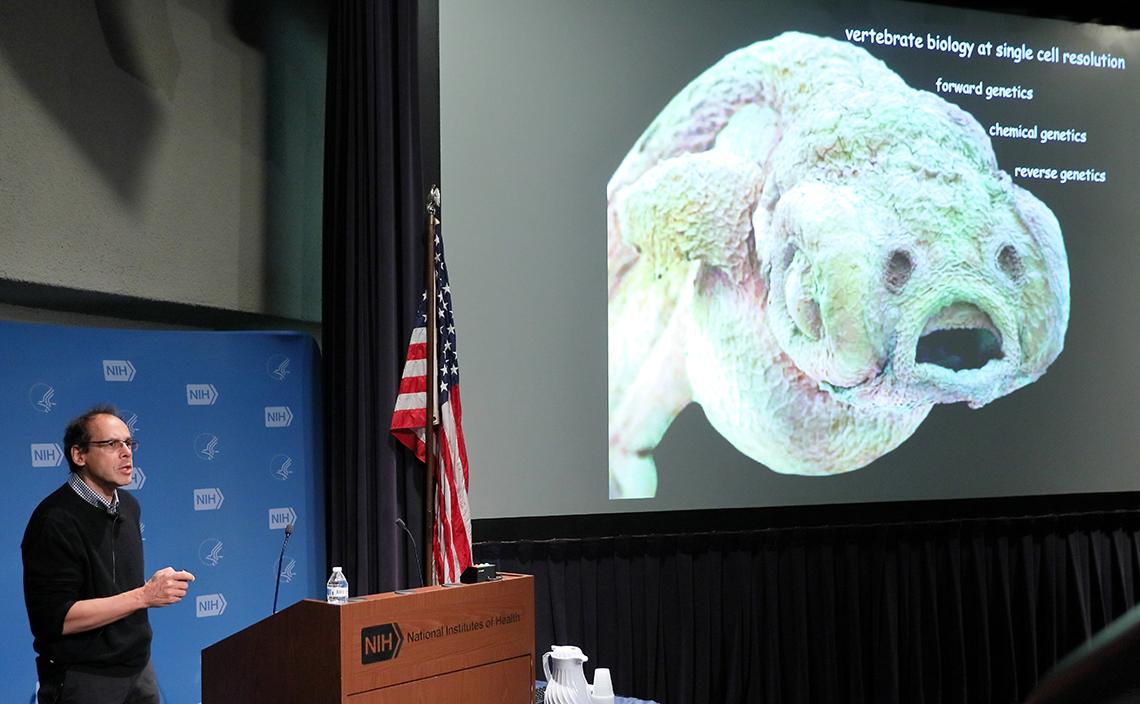 Stanier talking at the podium with a slide in the background.
