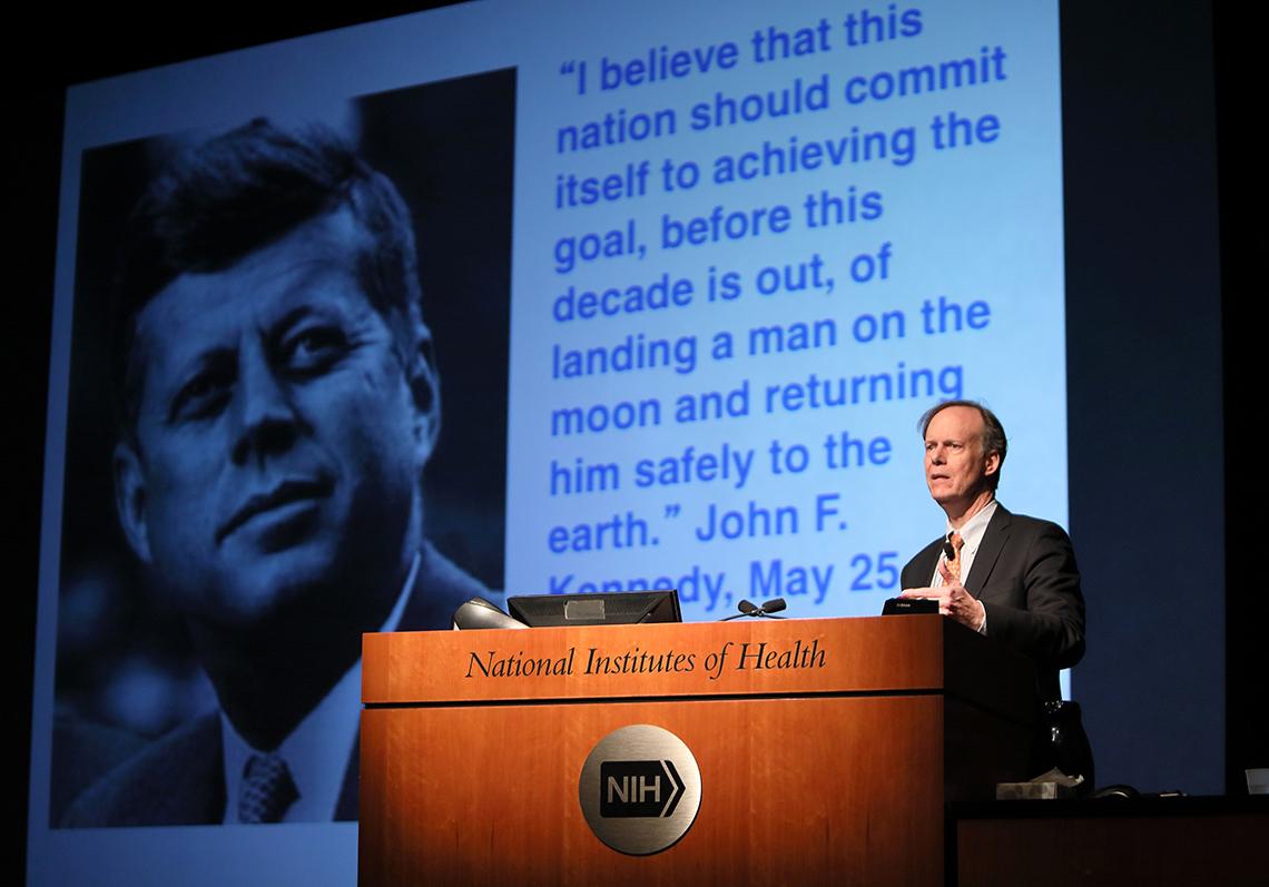 Kaelin speaking at the podium with a projected slide in the background.