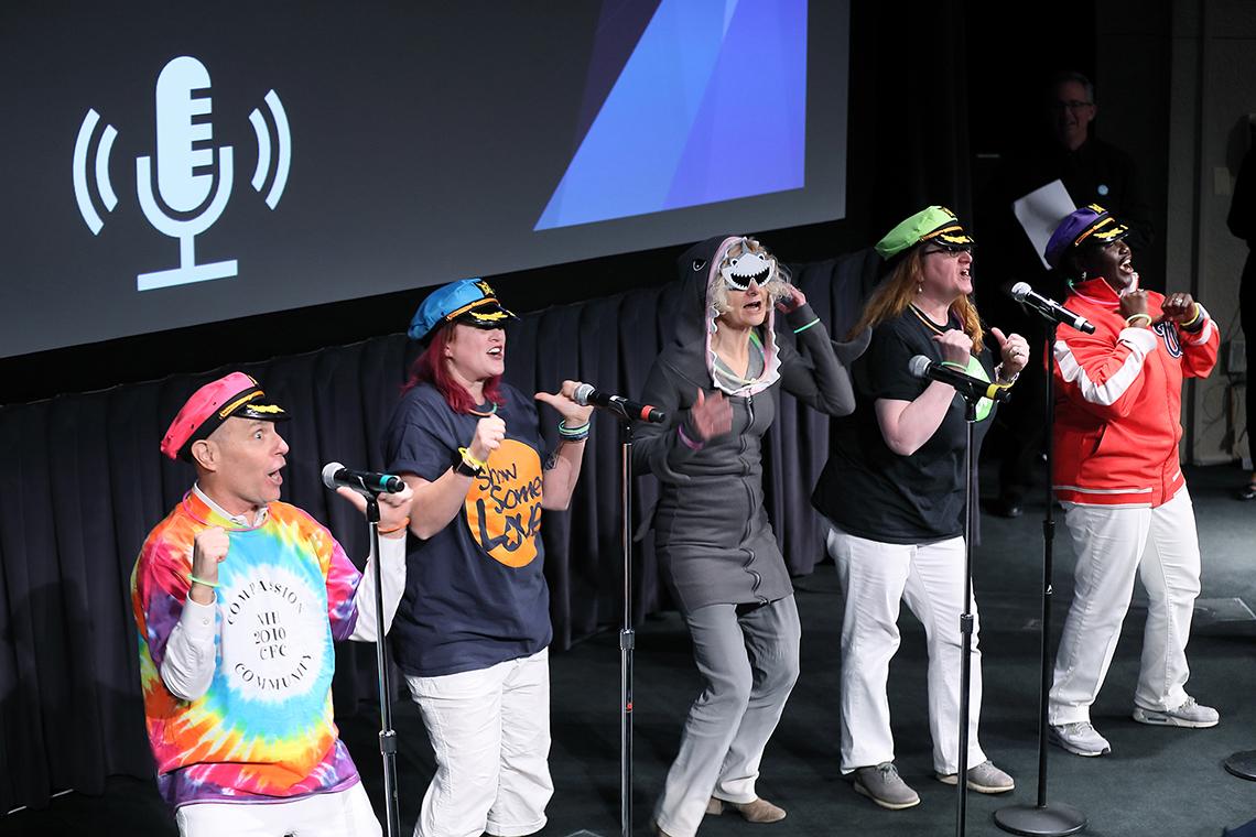 Five people, four wearing ship captain's hats and one wearing a shark costume, sing on stage.