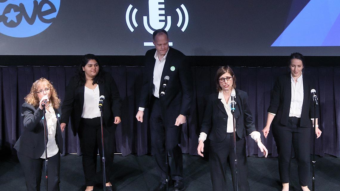 Five people wearing black suits with white shirts sing at the microphones