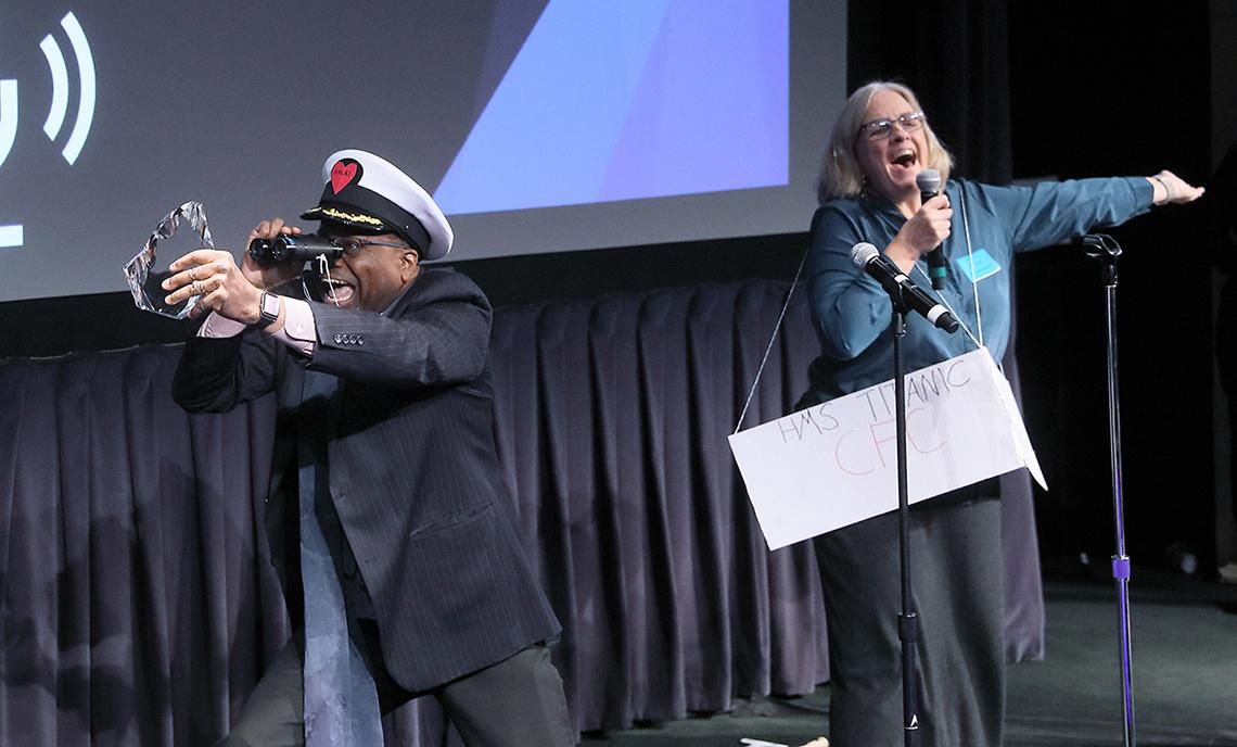 Gibbons wearing captain's hat peers through binoculars and holds up a glass paperweight as Dunbar sings