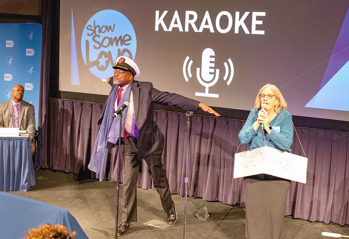Man in ship captain's hat mimics a masthead as woman with Titanic sign sings with mic.
