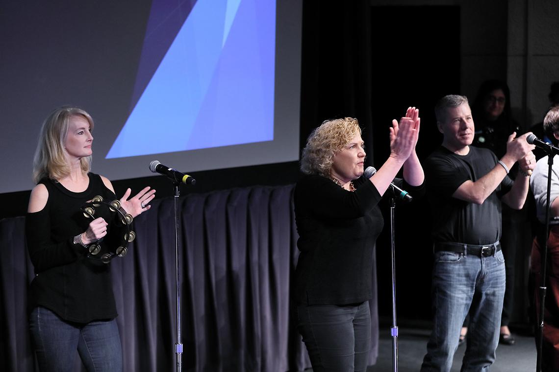 On stage, one woman plays tambourine, another claps, beside a man clapping.