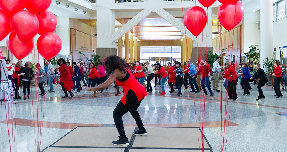 Fitness instructor leads a large group of NIH'ers in dance in Clinical Center atrium beneath red balloons.