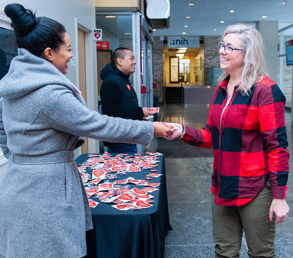 An NIH'er, beside a table of red heart pins, rewards a red shirt-wearing colleague with a pin in the Bldg. 31 lobby.