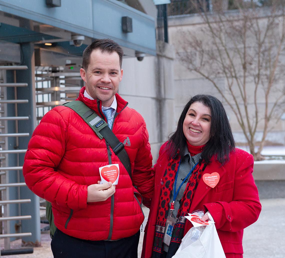 Two smiling NIH'ers in red jackets stand outside Metro station entrance eager to distribute heart stickers.