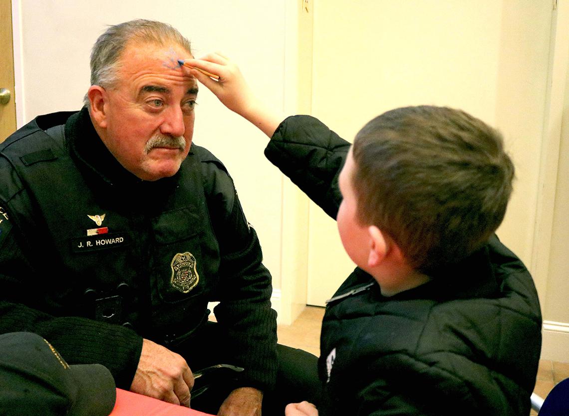A youngster paints the face of a police officer at a holiday party.