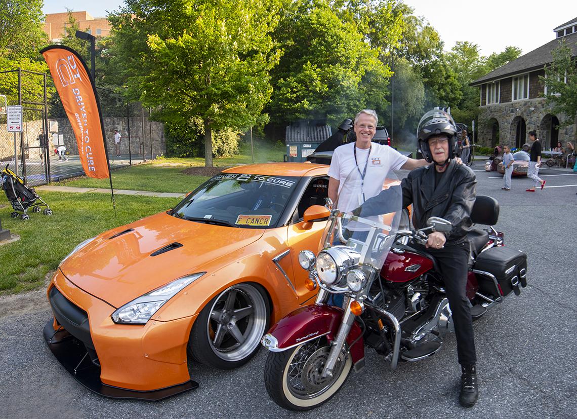 In front of Nissan GTR sportscar, Lee smiles with Collins on motorcycle