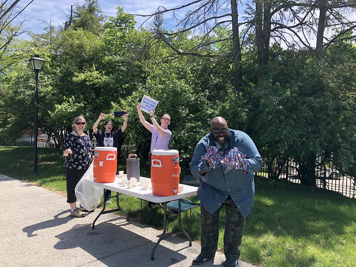 With water coolers on table, women cheer, wave signs, and man grins holding pompoms