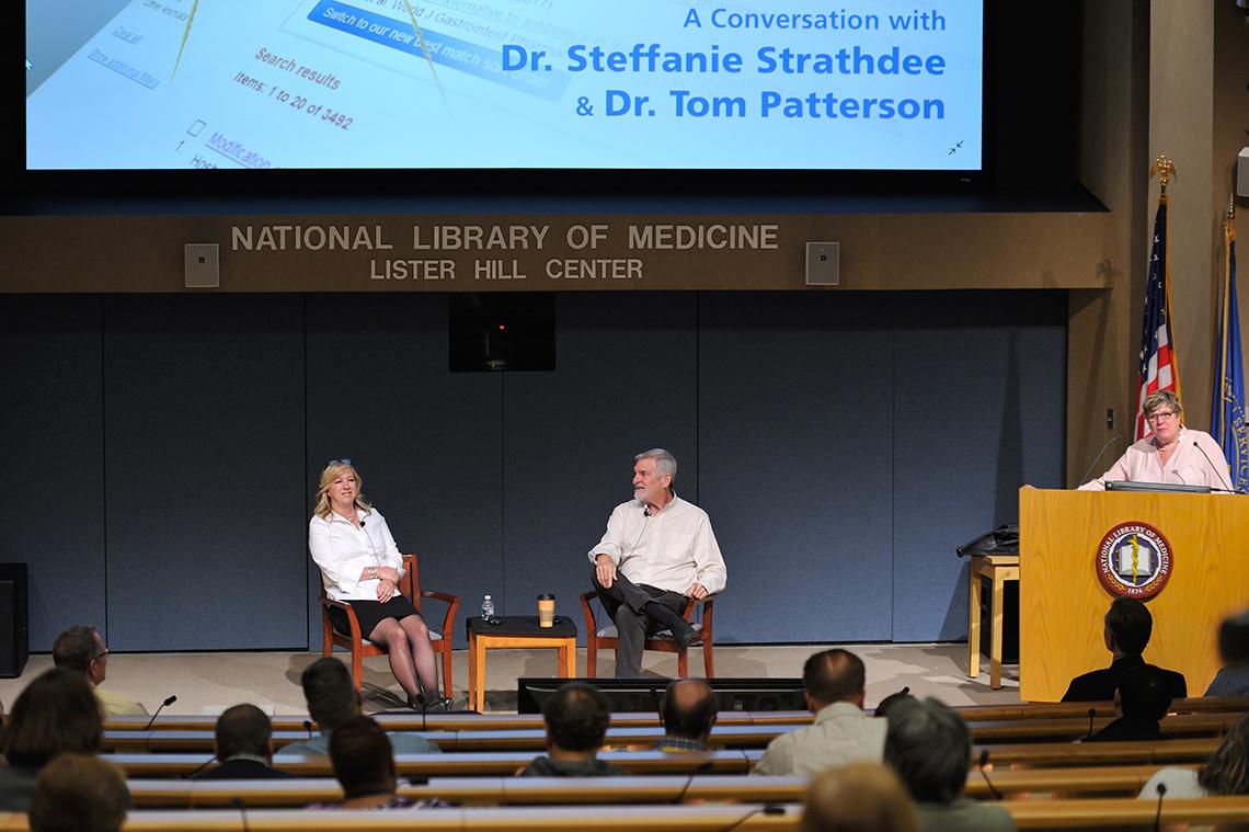 Brennan speaking at podium, with Strathdee and Patterson seated alongside on stage