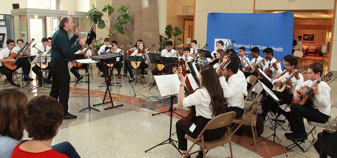 Male conductor gestures toward more than a dozen youngsters seated with their guitars