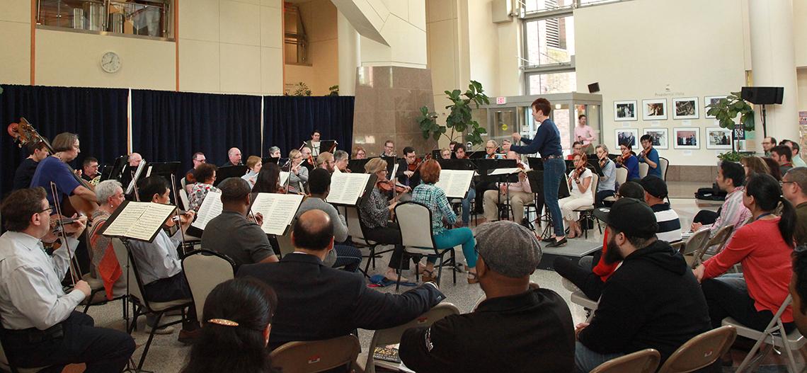 Dozens of musicians shown from behind, in the large, open atrium of the Clinical Center