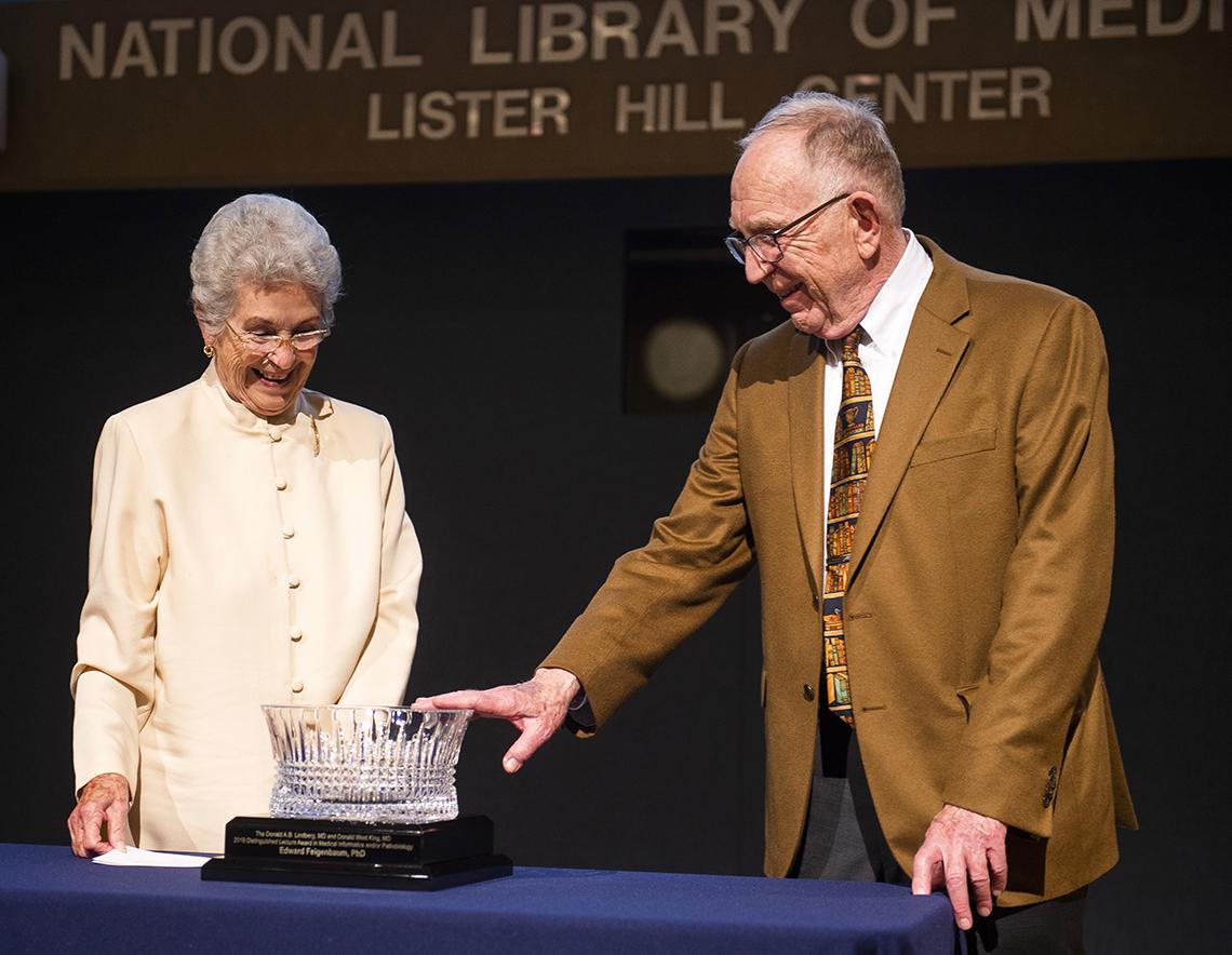 Mary Lindberg and Dr. Feigenbaum both smile as he admires, points to crystal bowl lecture award in Lister Hill Center.