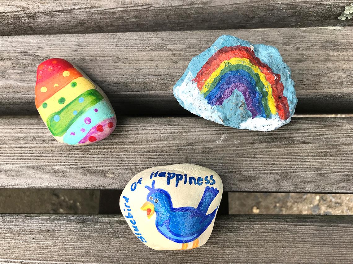 Three painted rocks on a bench, including a drawing of the bluebird of happiness.
