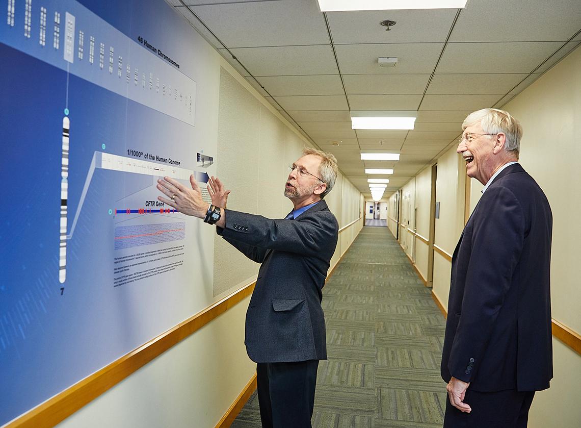 Dr. Green holds his hands up to wall of genome hallway exhibit as Dr. Collins marvels at the display.