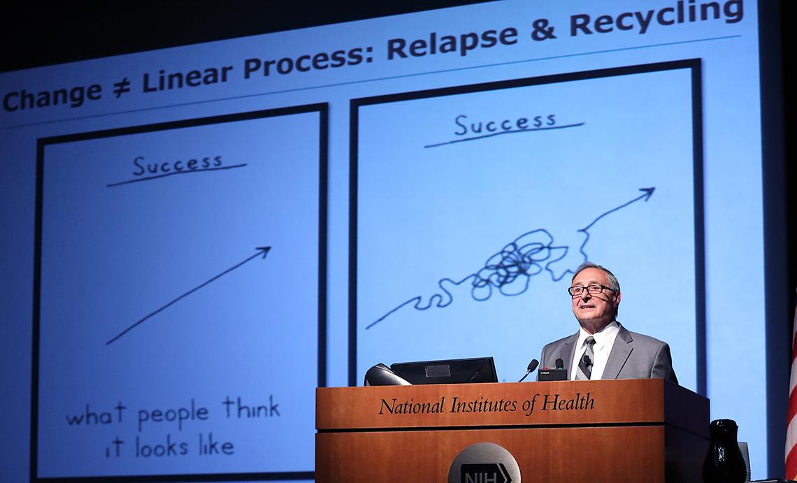 DiClimente talks in front of slide that shows squiggly line of conquering addiction, illustrating that success is not linear.