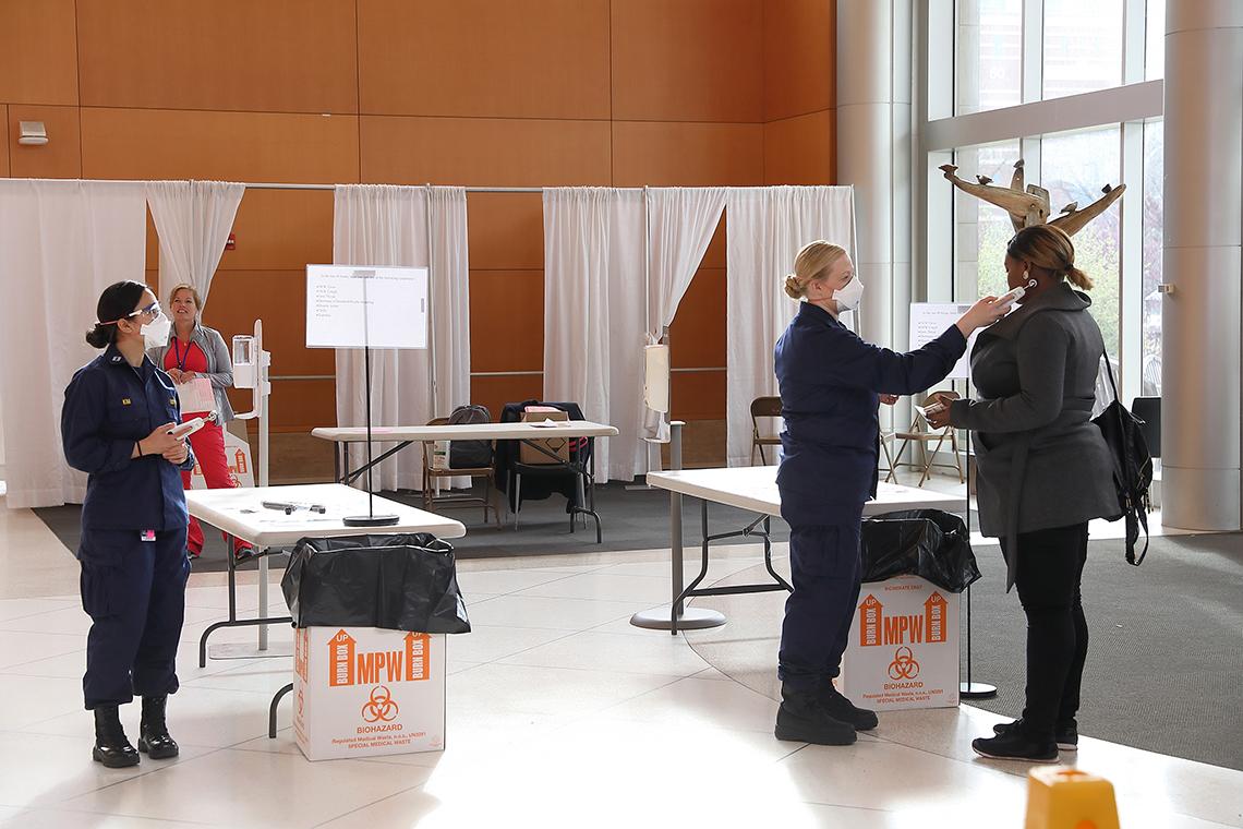 View of tables and makeshift screening areas as uniformed workers check temperatures of people entering building.