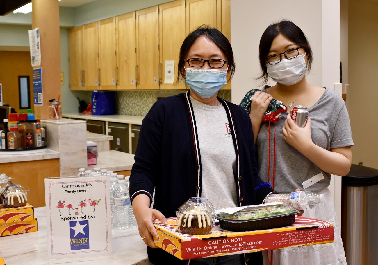 A young adult and her mom, smiling behind their masks, hold a pizza box, salad and cake.