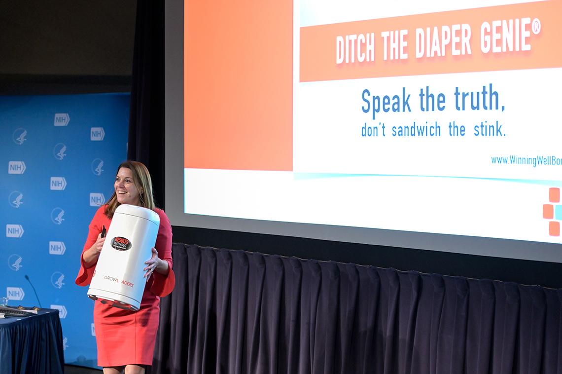 A smiling Karin Hurt walks across stage carrying a diaper pail, in front of a slide that reads, "Speak the Truth."