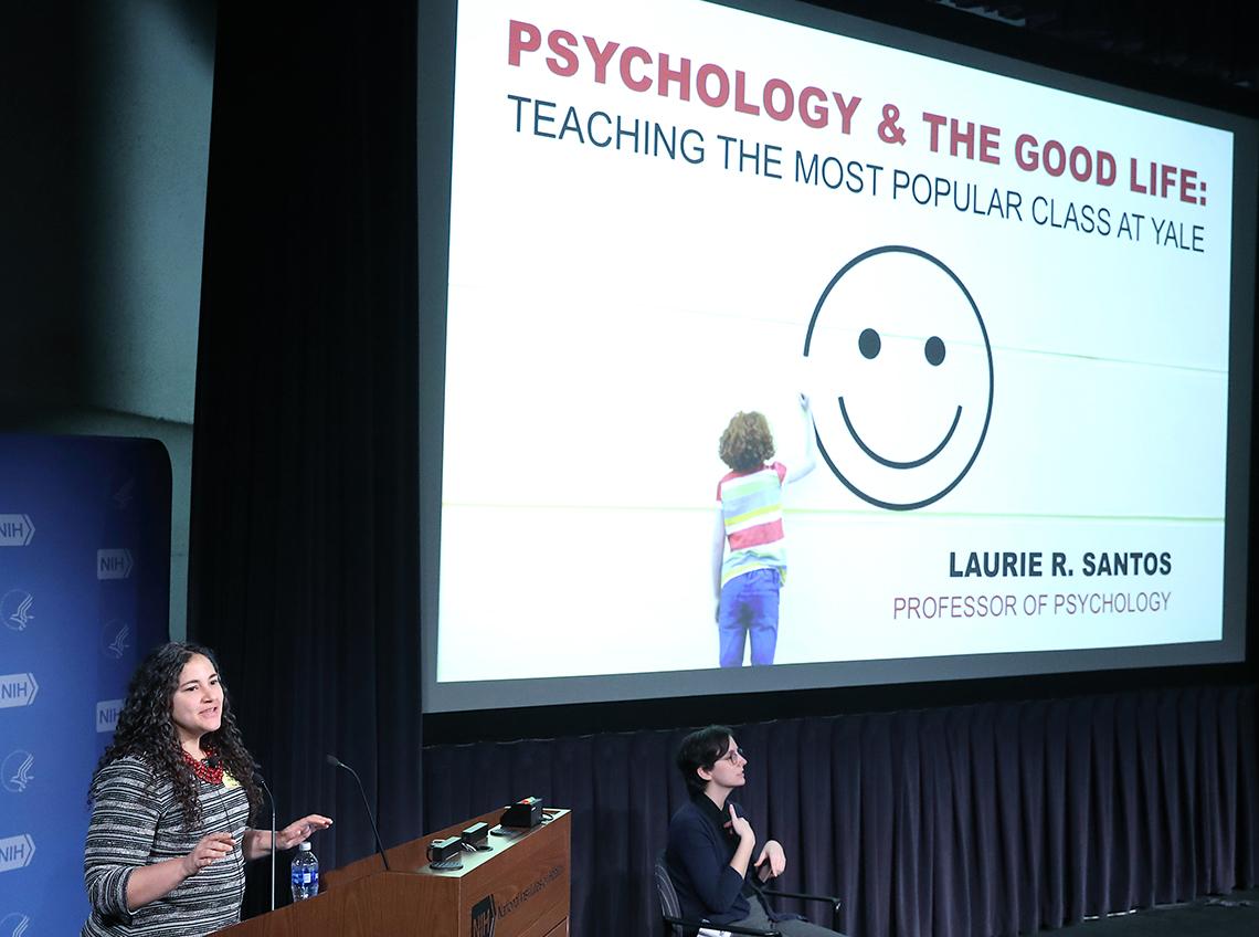 Santos stands in front of slide: Psychology and the Good Life, with a smiley face