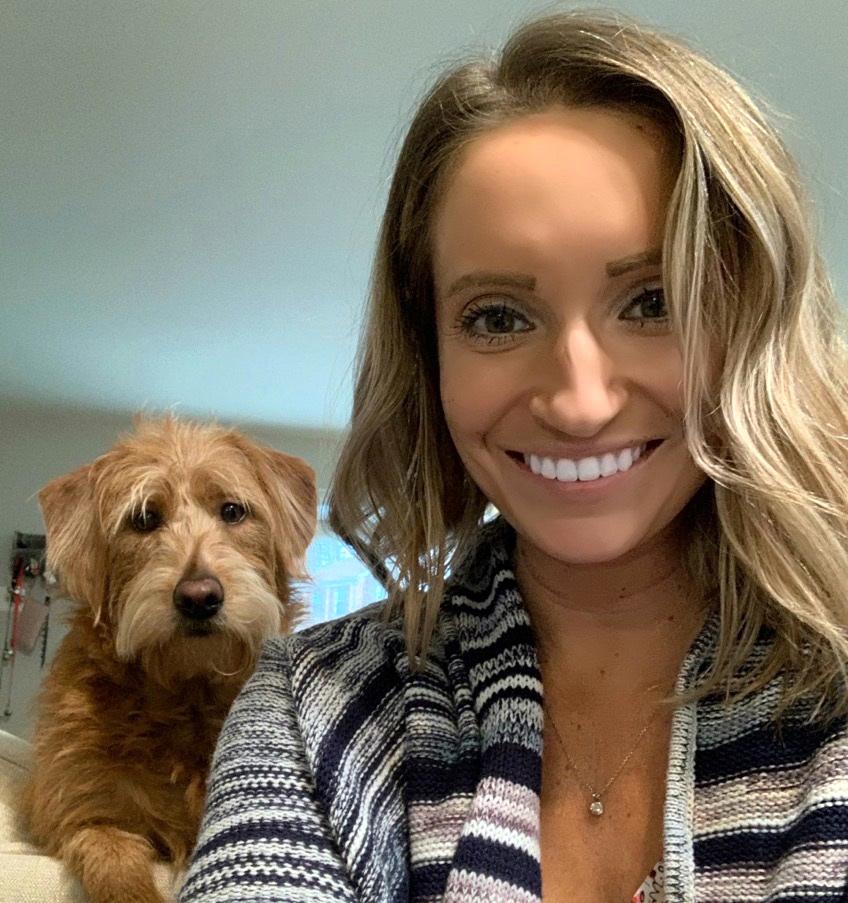 Dog looks over the shoulder of Alyssa Brooks as she smiles into the camera