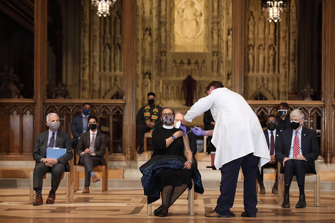 On the Cathedral stage, a seated nun receives an injection from a health worker in a lab coat.