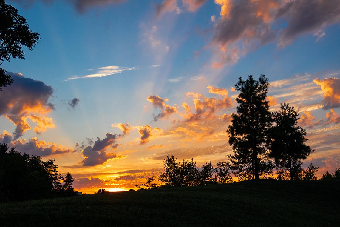 Yellow and peach surround clouds as the sun sets behind a field.