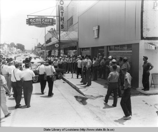 Black and white image of street corner where a group of people including police and photographers gather at a storefront.