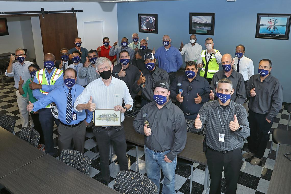 Collins holds up image of the jet engine and signals thumbs up with large group of CUP staff--all in face masks-- joining him.