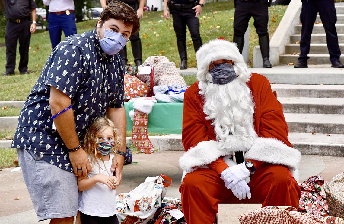 A man and his daughter stand next to seated, masked Santa.