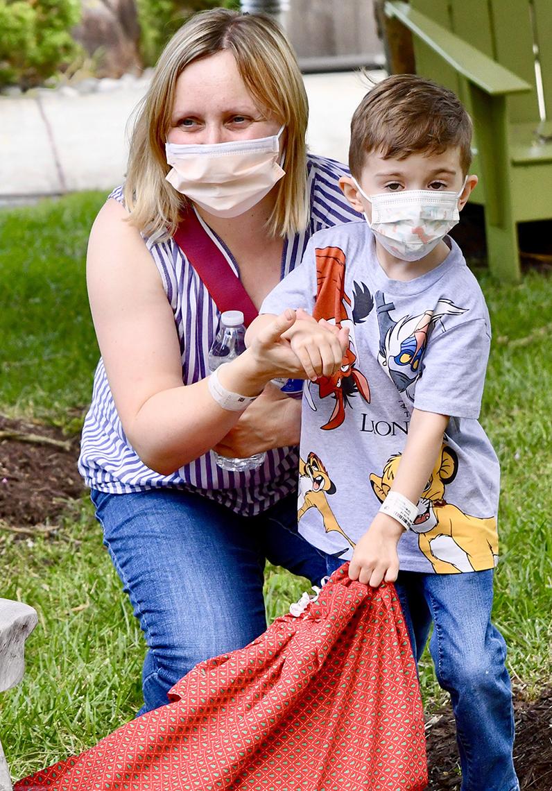 A woman stoops next to a little boy and holds his hand while he holds a bag. 