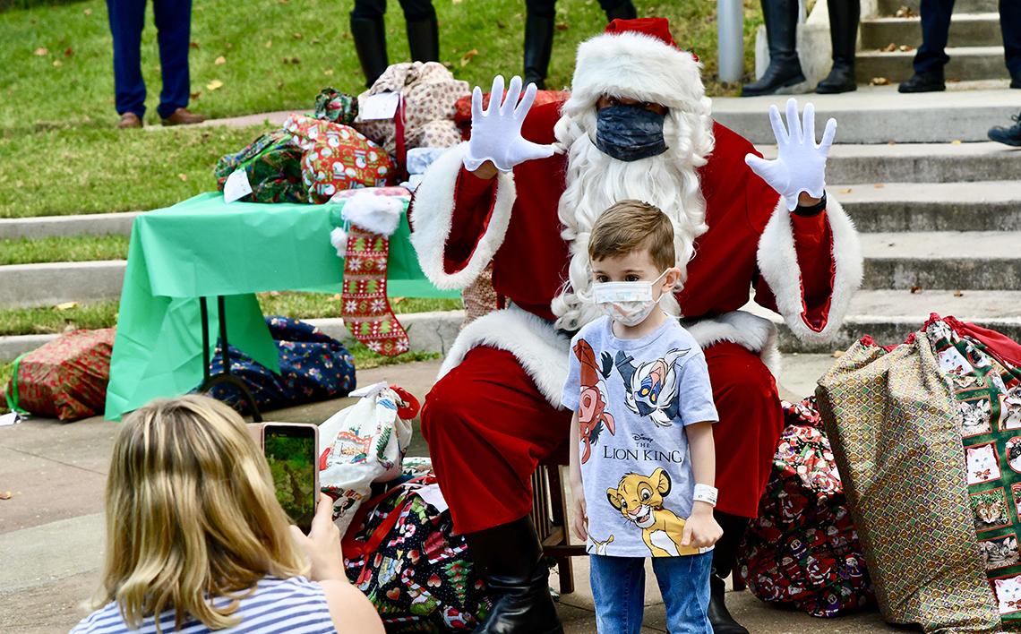 A little boy stands in front of masked Santa, who is holding his hands up with open palms 