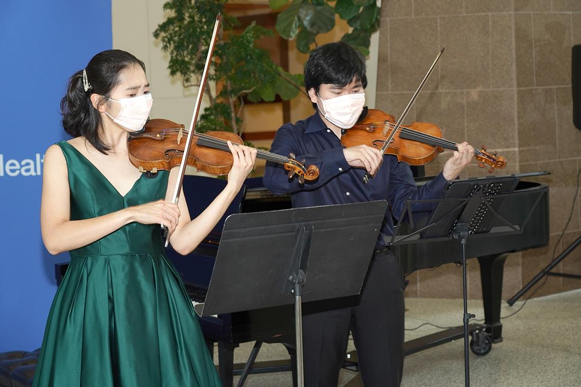 Two young adults play violins while masked in the atrium of the CC